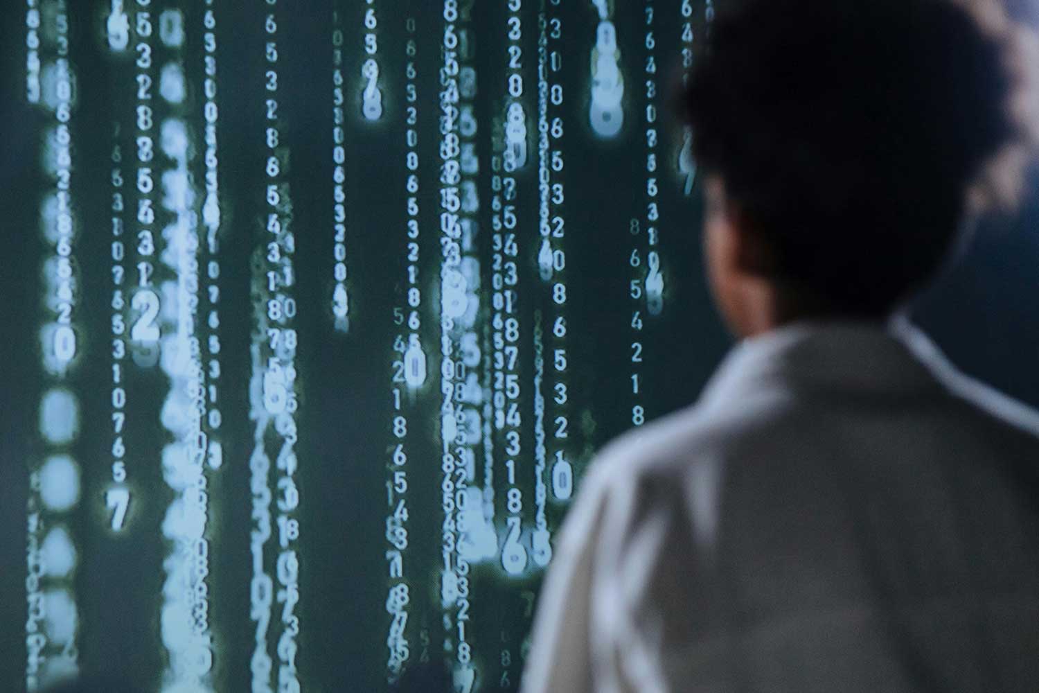 Person von hinten betrachtet einen Bildschirm mit vertikalen Reihen aus leuchtenden Zahlen und Binärcode, ähnlich einer Matrix-Darstellung.