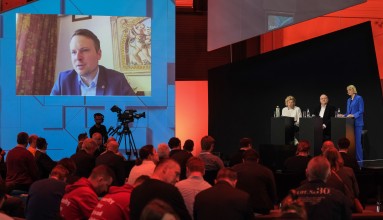 Publikum vor einer Bühne mit drei Rednern