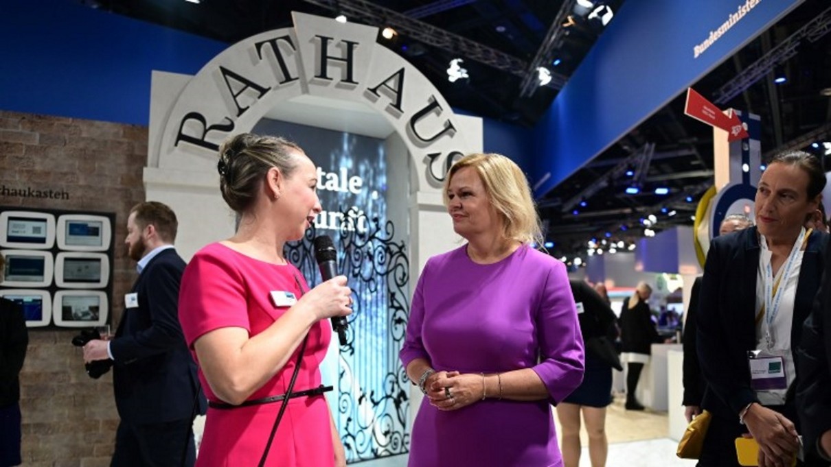 Two women in the foreground, Federal Minister of the Interior Nancy Faeser on the BMI stand on the right, the backdrop of a town hall gate in the background.