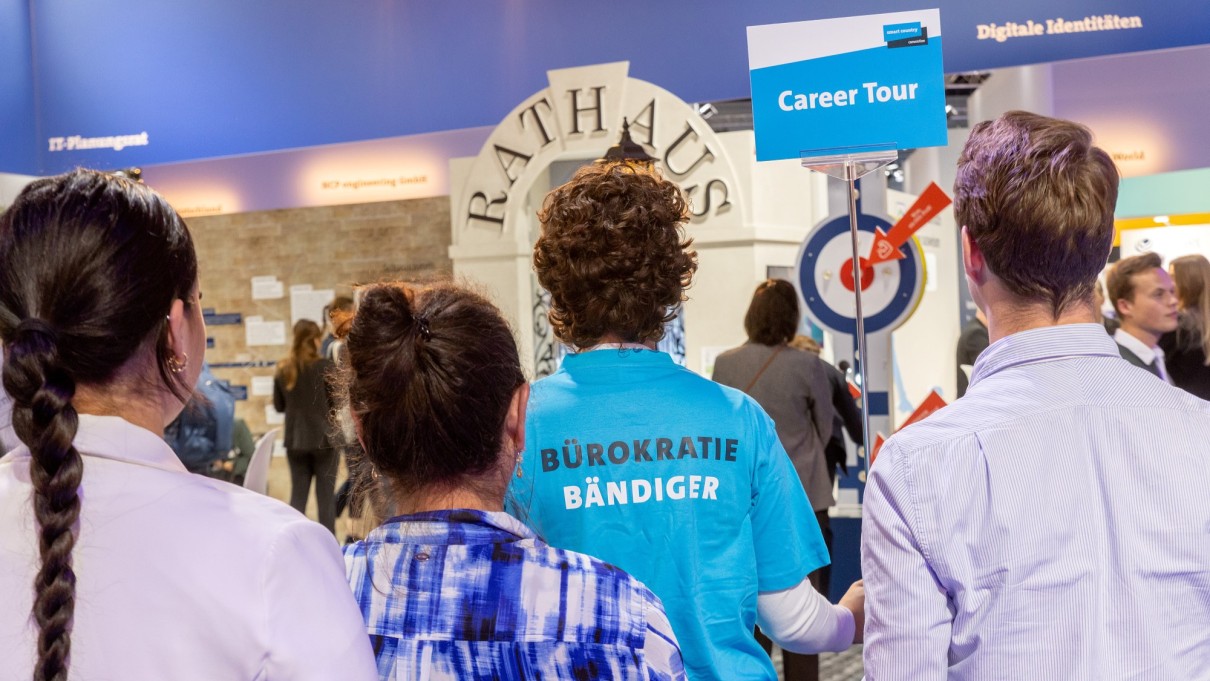 Four people from the back of the booth of the Federal Ministry of the Interior and Homeland, one person holds a sign with the inscription 'Career Tour'.