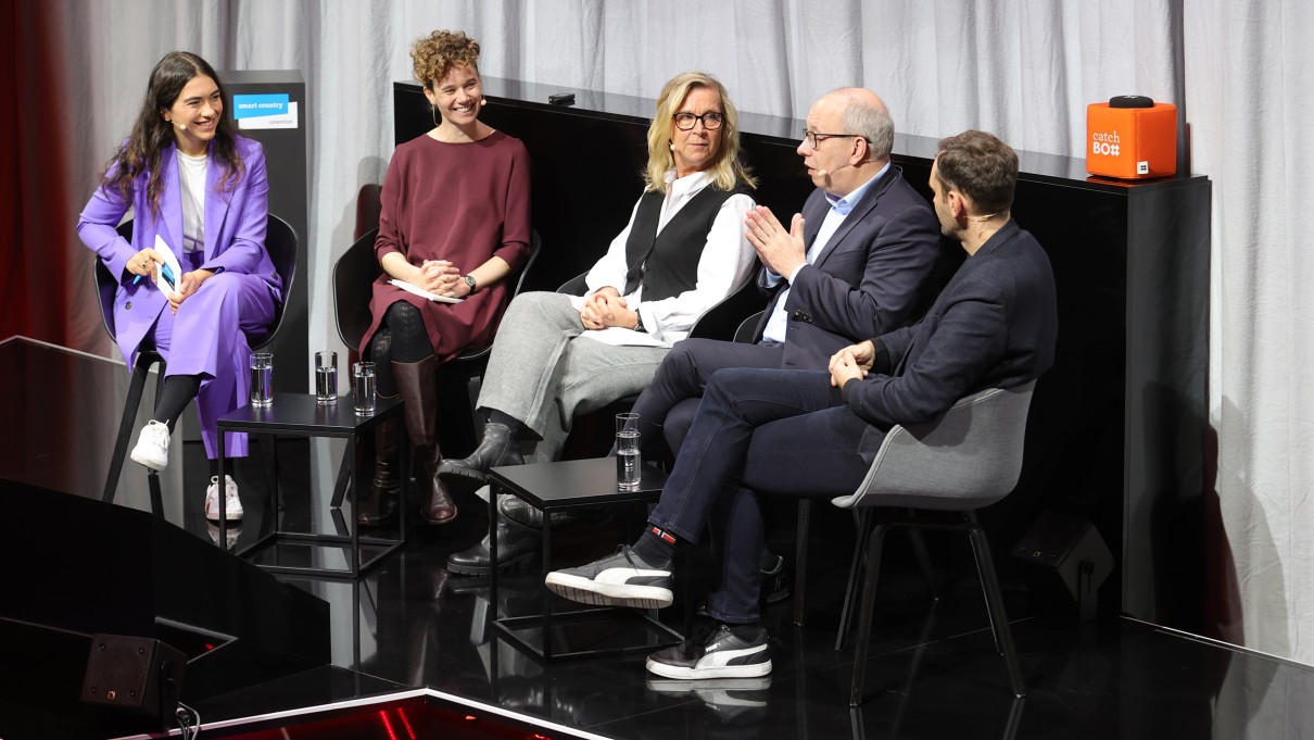 Auf einer Bühne sitzen eine Moderatorin und vier Panelistinnen und Panelisten, die miteinander im Gespräch sind. Auf einer Bühne sitzen eine Moderatorin und vier Panelistinnen und Panelisten, die miteinander im Gespräch sind.