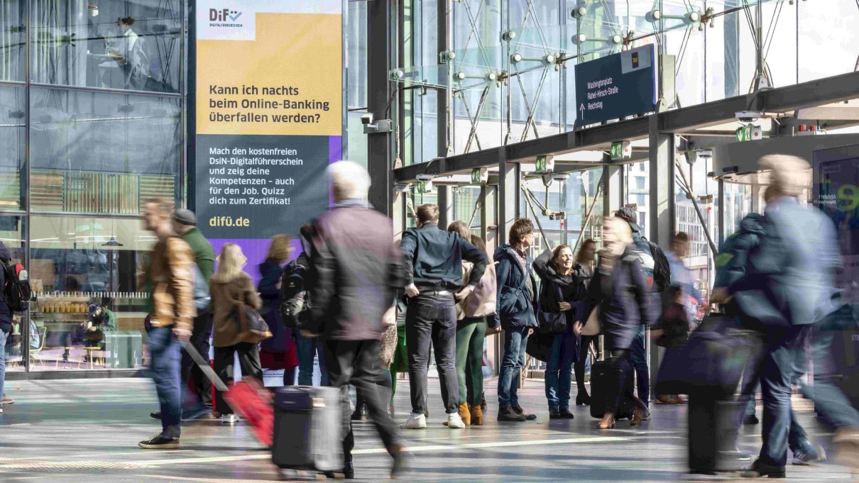 Menschen am Bahnhof, im Hintergrund eine großflächige Werbung zum DiFü Menschen am Bahnhof, im Hintergrund eine großflächige Werbung zum DiFü