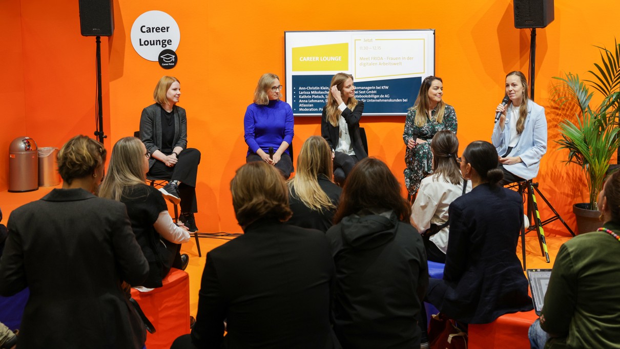 Auf einer Bühne sitzen eine Moderatorin und vier Panelistinnen, die miteinander im Gespräch sind. Im Vordergrund das Publikum. Auf einer Bühne sitzen eine Moderatorin und vier Panelistinnen, die miteinander im Gespräch sind. Im Vordergrund das Publikum.