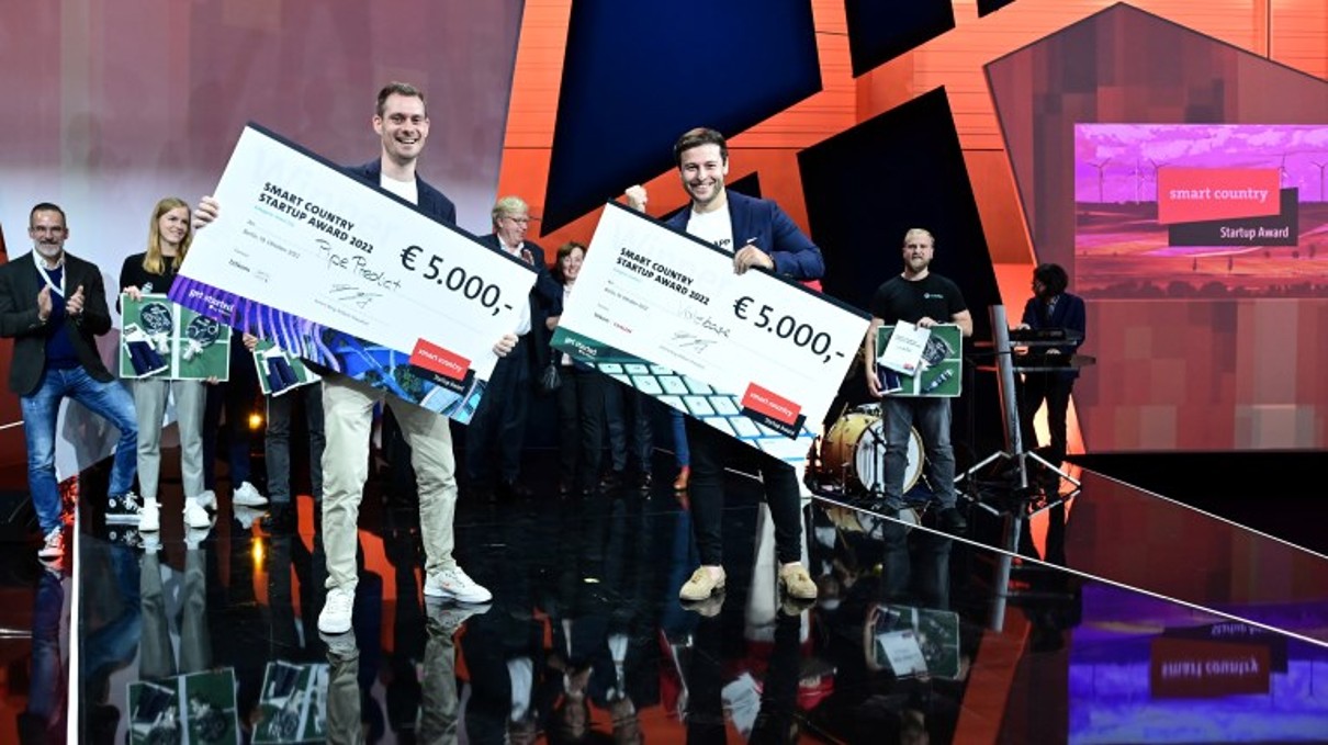 Auf der Bühne stehen zwei Männer stolz und präsentieren die großen Schecks mit den 5.000 Euro Preisgeld in ihren Händen. Auf der Bühne stehen zwei Männer stolz und präsentieren die großen Schecks mit den 5.000 Euro Preisgeld in ihren Händen.