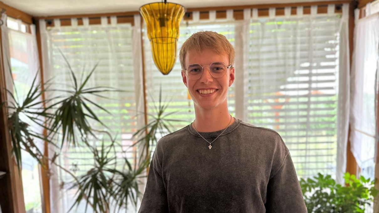 Young man in the foreground, large windows with blinds and green plants in the background.