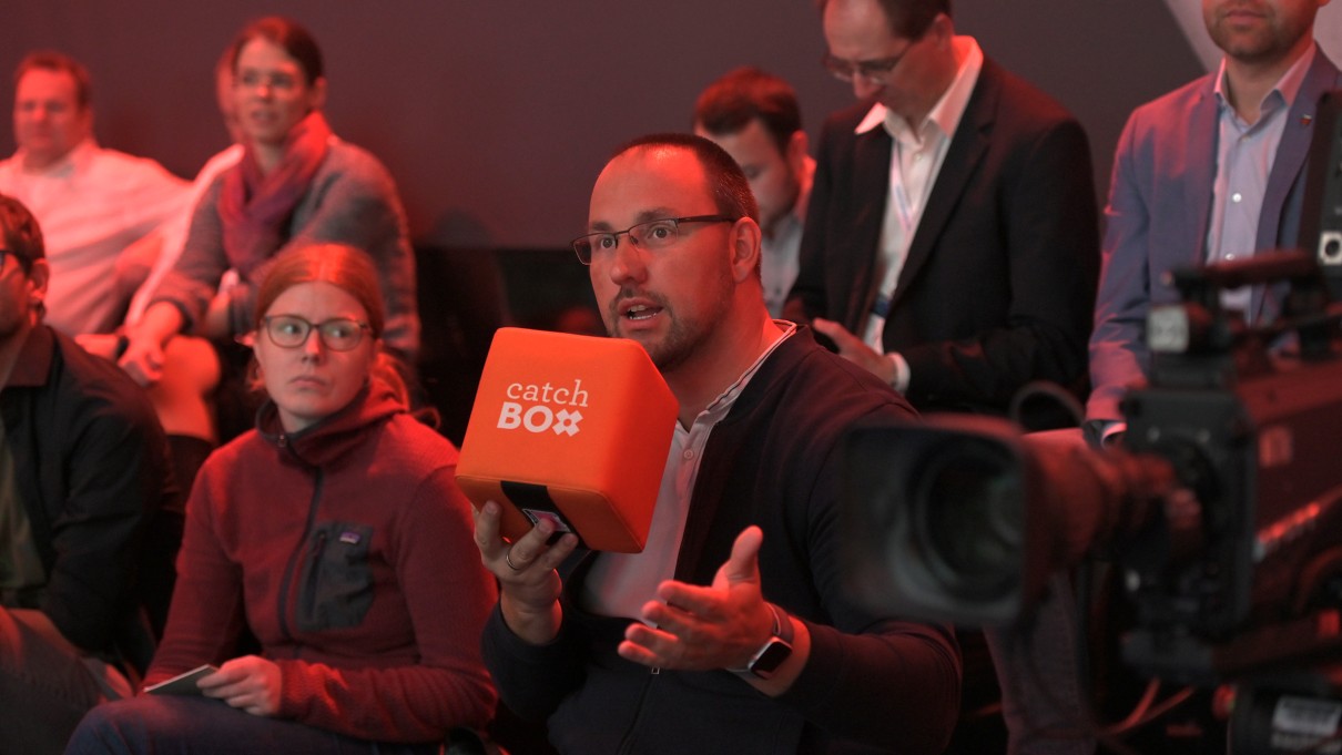 Eine Gruppe von Zuschauerinnen und Zuschauern ist abgebildet. Ein Mann hält ein Mikrofon in der Hand. Eine Gruppe von Zuschauerinnen und Zuschauern ist abgebildet. Ein Mann hält ein Mikrofon in der Hand.