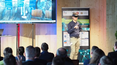 Ein Mann präsentiert eine Virtual Reality Brille, im Vordergrund ist das Publikum von hinten zu sehen