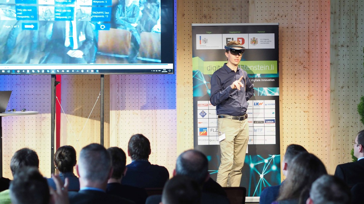 Ein Mann präsentiert eine Virtual Reality Brille, im Vordergrund ist das Publikum von hinten zu sehen. Ein Mann präsentiert eine Virtual Reality Brille, im Vordergrund ist das Publikum von hinten zu sehen.