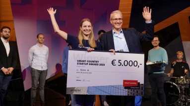 Sieger des Smart Country Startup Awards SUMM AI auf der SCCON Bühne, ein Mann und eine Frau mit einem großen Scheck in der Hand.
