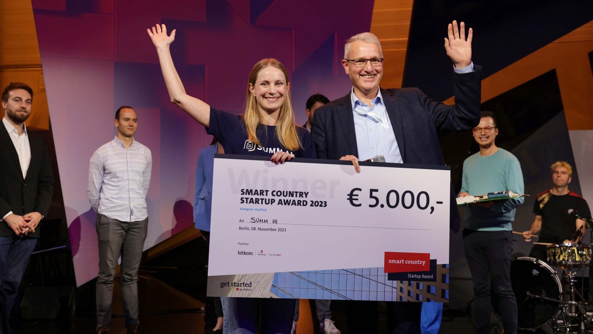 Sieger des Smart Country Startup Awards SUMM AI auf der SCCON Bühne, ein Mann und eine Frau mit einem großen Scheck in der Hand. Im Hintergrund sind die anderen Finalisten zu sehen. Sieger des Smart Country Startup Awards SUMM AI auf der SCCON Bühne, ein Mann und eine Frau mit einem großen Scheck in der Hand. Im Hintergrund sind die anderen Finalisten zu sehen.