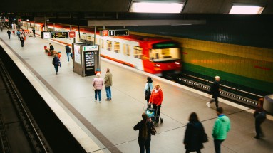 Bahnsteig mit einfahrender Bahn, Menschen im Vordergrund. 