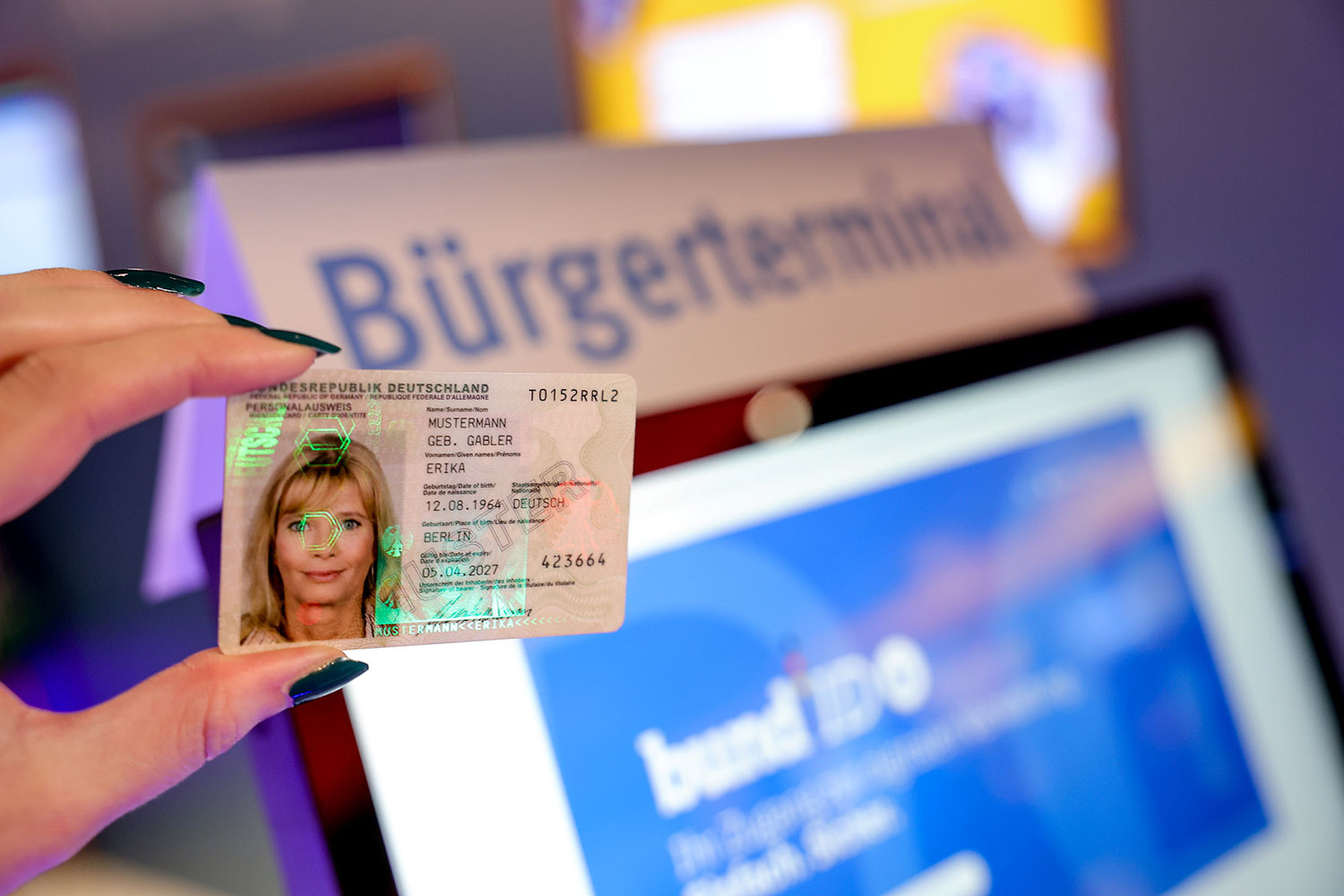 Im Vordergrund eine Hand, die einen Personalausweis hält. Im Hintergrund ein Bürgerterminal zur BundID. Im Vordergrund eine Hand, die einen Personalausweis hält. Im Hintergrund ein Bürgerterminal zur BundID.